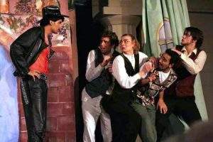 Vor dem Bühnenbild einer Mauer mit Durchgang steht ein in Schwarz und Rot gekleideter jugendlicher Schauspieler in selbstbewusster Pose. 4 andere jugendliche Schauspieler ziehen sich sichtlich erschrocken zurück.