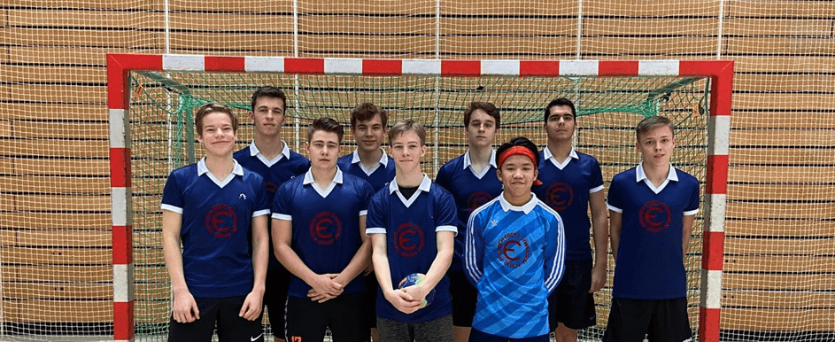 Auf einem Gruppenfoto vor dem Handballtor stehen 9 Mannschaftsmitglieder der FEO-Jungen-Mannschaft. Sie tragen ihre blauen Mannschaftstrikots und schwarze Sportshorts und schauen lächelnd in die Kamera.