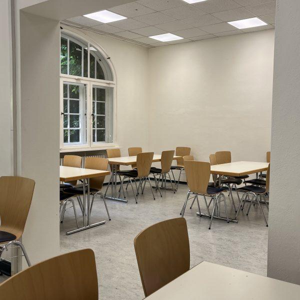 Zwei durch einen großen Durchbruch miteinander verbundene Räume: Weiße Wände, moderne weiße Tische und Stühle aus hellem Holz mit dunkelgrau gepolsterten Sitzflächen.