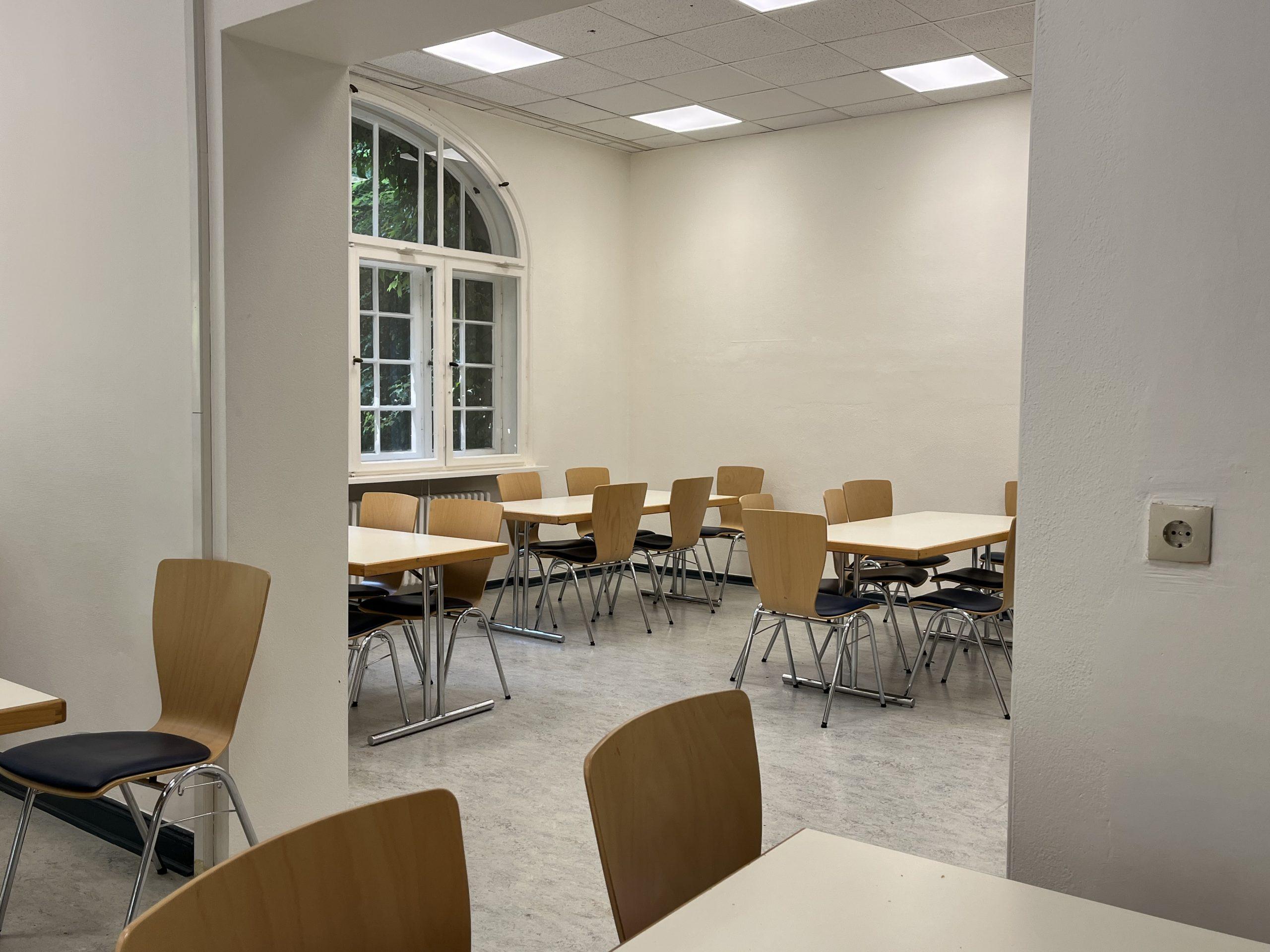 Zwei durch einen großen Durchbruch miteinander verbundene Räume: Weiße Wände, moderne weiße Tische und Stühle aus hellem Holz mit dunkelgrau gepolsterten Sitzflächen.