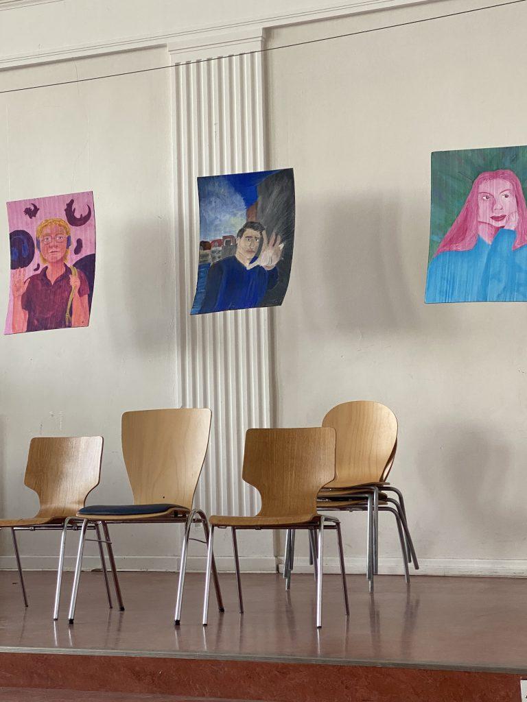 Hinter eine Reihe aus Stühlen an der Wand der Aula 3 gemalte Porträtbilder: Eine Frau trägt Kopfhörer, ein Mann streckt die Hand abwehrend nach vorn, eine andere Frau stützt ihren Kopf auf die Hand