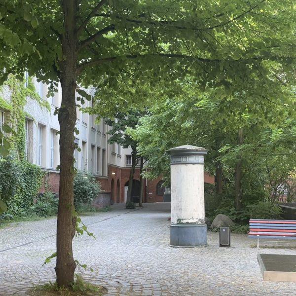 Der grau gepflasterte Schulhof mit vielen Bäumen. Eine altmodische weiße Litfaßsäule, eine rot-blau-gestrichenen Holzbank und eine Tischtennisplatte sind zu sehen.