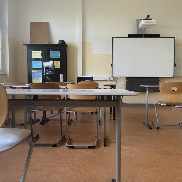 Klassenraum mit hellen Holzstühlen und weißen Tischen. An der gelben Wand ein Activboard, daneben ein Whiteboard. Auf dem Lehrertisch ein Laptop.