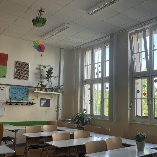 Im hellen Raum stehen Schulbänke mit weißen Tischplatten und Stühle aus hellem Holz. An den Fenstern kleben bunte handtellergroße Papierkreise. An den Wänden bunte Plakate. Mehrere Zimmerpflanzen stehen im Raum.