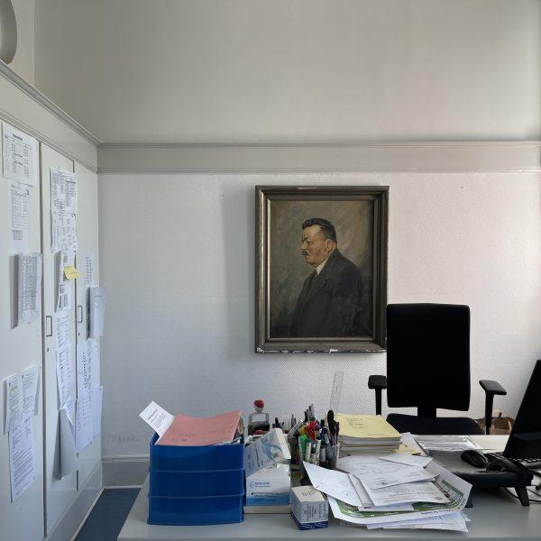 An der Wand hängt ein Friedrich-Ebert-Porträt in altem Holzrahmen. Auf dem Schreibtisch ein großer Flachbildschirm, eine Tastatur und ein Telefon sowie viele Schreibutensilien und mehrere Dokumenten-Stapel. Am weißen Einbauschrank links hängen eine Uhr und viele Listen.