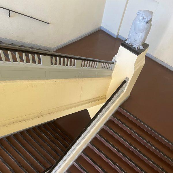 Das hellgelb gestrichene Treppenhaus mit braunem Linoleum und gedrechseltem Treppengeländer. Eine große graue Eulenstatue ziert den Geländer-Eckpfosten.